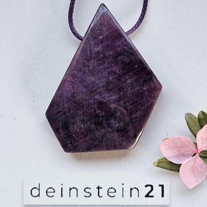 Könnte beinhalten: Ein facettierter, tiefvioletter Edelsteinanhänger mit geometrischer Form, an einer passenden Kordel. Der Anhänger befindet sich vor einem weißen Hintergrund, mit dem Text "deinstein21". Eine rosa Blume mit grünen Blättern ist in der Ecke.