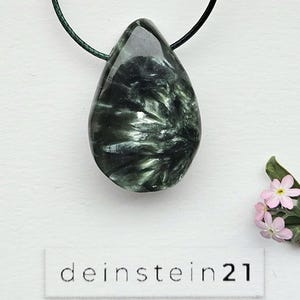 Può includere: Collana con pendente a goccia in pietra preziosa verde scuro lucida con motivo a vortice. Il pendente è sospeso a un cordino verde scuro. Il testo "deinstein21" è stampato su un'etichetta bianca sotto il pendente.