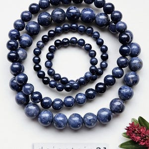 Può includere: Quattro collane di perle di pietre preziose blu di diverse lunghezze. Le collane sono fatte di perle rotonde e sono disposte in cerchio. Le perle sono di un blu profondo con un leggero luccichio. Le collane sono su uno sfondo bianco.