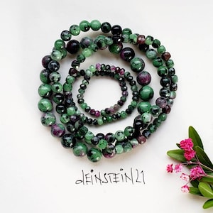 Könnte beinhalten: Ein Set aus vier grünen und violetten Edelstein-Armbändern. Die Armbänder bestehen aus runden Perlen und sind übereinander gestapelt. Der Text "deinSTEIN/21" ist auf einer weißen Oberfläche unter den Armbändern geschrieben.