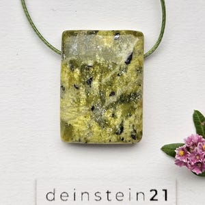 Pode incluir: Um pingente retangular com um cordão verde. O pingente é de cor verde claro com manchas pretas. O texto "deinstein21" está impresso em uma etiqueta branca abaixo. Um pequeno grupo de flores cor-de-rosa está no canto inferior direito.