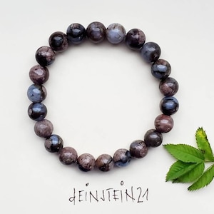 Könnte beinhalten: Ein Armband aus dunkelblauen, violetten und grauen Edelsteinperlen. Das Armband liegt auf einem weißen Hintergrund mit einer handschriftlichen Signatur, die "deinJTEIN21" lautet.