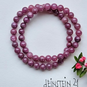 Könnte beinhalten: Ein doppelreihiges Armband aus rosafarbenen und violetten Edelsteinperlen. Das Armband liegt auf einer weißen Oberfläche mit dem Text "deinSTEIN 21" darunter.