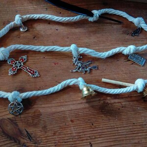 May include: A white rope adorned with various charms: a cross, a witch on a broom, a pentagram, a bell, and a miniature broom. The charms are silver and red, displayed on a wooden surface.