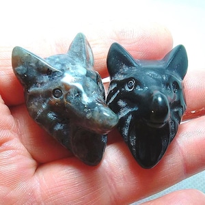 May include: Two carved stone wolf head figurines. One is a mottled grey and brown, the other is solid black. The wolf heads have pointed ears, detailed noses, and are held in a hand.