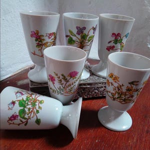 May include: A collection of six white porcelain goblets, each adorned with a unique floral pattern. The goblets showcase botanical illustrations in pink, purple, yellow, and green. They have a flared design and are supported by a pedestal base.