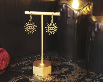 Puede incluir: Pendientes de tono dorado con un diseño de sol y ojo. Los pendientes se muestran en un soporte dorado sobre un fondo negro.