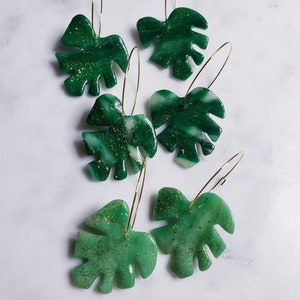 May include: Five pairs of green and white marbled leaf-shaped earrings with gold hoops. The leaves have a glittery finish.