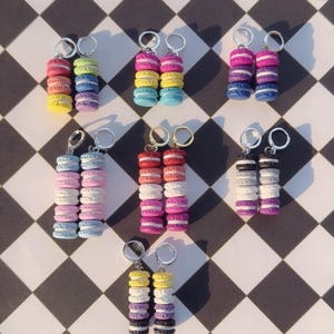 Könnte beinhalten: Eine Sammlung bunter Macaron-Ohrringe. Jedes Paar besteht aus einem Stapel winziger, farbenfroher Macarons, darunter Rosa, Gelb, Blau und Lila. Die Ohrringe werden auf einer schwarz-weiß karierten Oberfläche präsentiert.