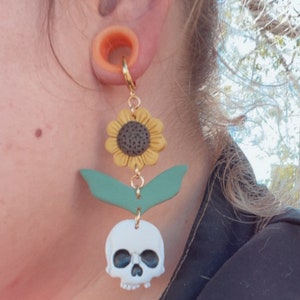May include: A gold-colored hoop earring with a sunflower, green leaves, and a white skull charm. The sunflower has yellow petals and a dark center. The skull has black eye sockets. The earring is attached to a flesh-colored ear tunnel.