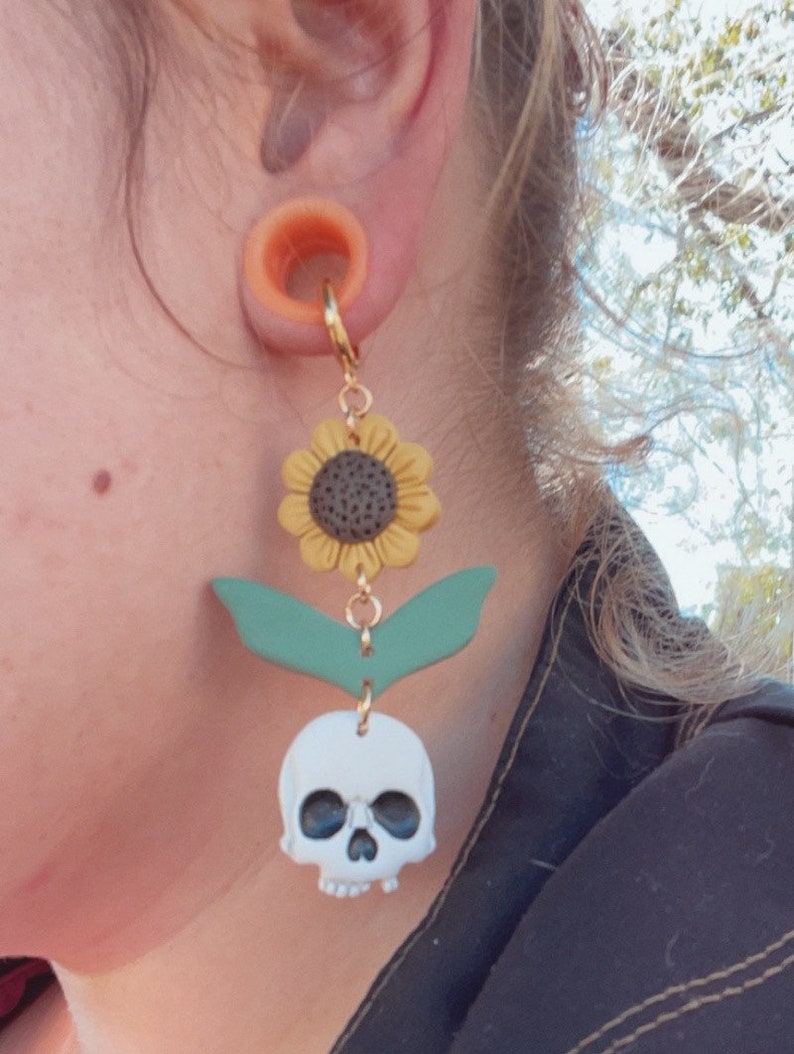 May include: A gold-colored hoop earring with a sunflower, green leaves, and a white skull charm. The sunflower has yellow petals and a dark center. The skull has black eye sockets. The earring is attached to a flesh-colored ear tunnel.