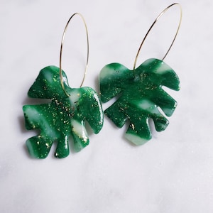 May include: A pair of green and white swirl patterned earrings with gold glitter accents. The earrings are shaped like monstera leaves and hang from gold hoop earrings.