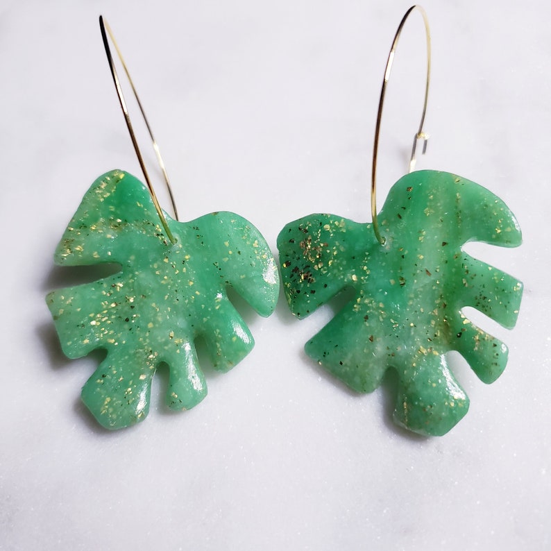 May include: A pair of green and gold glitter monstera leaf earrings with gold hoop earrings.