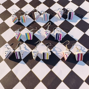 May include: A selection of cake-shaped earrings in various designs, including rainbow, pink, purple, and monochrome. Each earring has a silver-coloured hook. The earrings are displayed on a black and white chequered surface. The earrings are a fun accessory.
