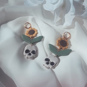 May include: A pair of handmade earrings featuring a skull, sunflower, and green leaf design. The earrings have a gold-colored hoop and are made of polymer clay. The skull is light gray with black eye sockets and a jawline. The sunflower is yellow and brown.