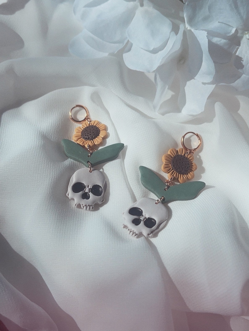 May include: A pair of handmade earrings featuring a skull, sunflower, and green leaf design. The earrings have a gold-colored hoop and are made of polymer clay. The skull is light gray with black eye sockets and a jawline. The sunflower is yellow and brown.