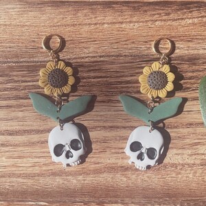 May include: A pair of dangle earrings featuring a sunflower, green leaves, and a skull. The earrings have gold-colored hoops and are set against a wooden background. The sunflowers are yellow and brown, and the skulls are white.