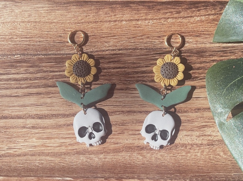 May include: A pair of dangle earrings featuring a sunflower, green leaves, and a skull. The earrings have gold-colored hoops and are set against a wooden background. The sunflowers are yellow and brown, and the skulls are white.