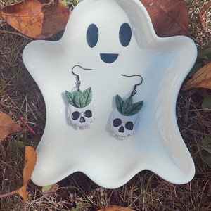 Puede incluir: Pendientes con forma de calavera con detalles de hojas verdes, presentados en un plato blanco con forma de fantasma. Los pendientes tienen ganchos negros. El plato est&aacute; sobre hierba y hojas ca&iacute;das, creando un tema de Halloween.