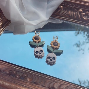 May include: A pair of unique earrings featuring a skull, sunflower, and leaf design. The earrings have a gray skull pendant, a yellow and brown sunflower, and green leaves. The earrings are displayed on a reflective surface.