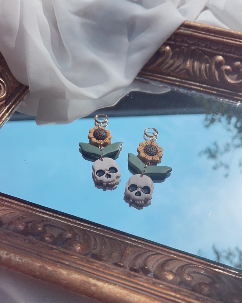 May include: A pair of unique earrings featuring a skull, sunflower, and leaf design. The earrings have a gray skull pendant, a yellow and brown sunflower, and green leaves. The earrings are displayed on a reflective surface.