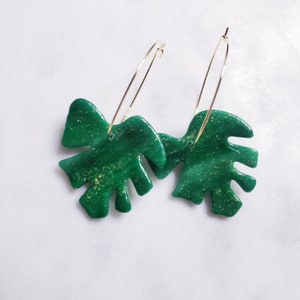 May include: A pair of gold hoop earrings with green glitter monstera leaf charms.