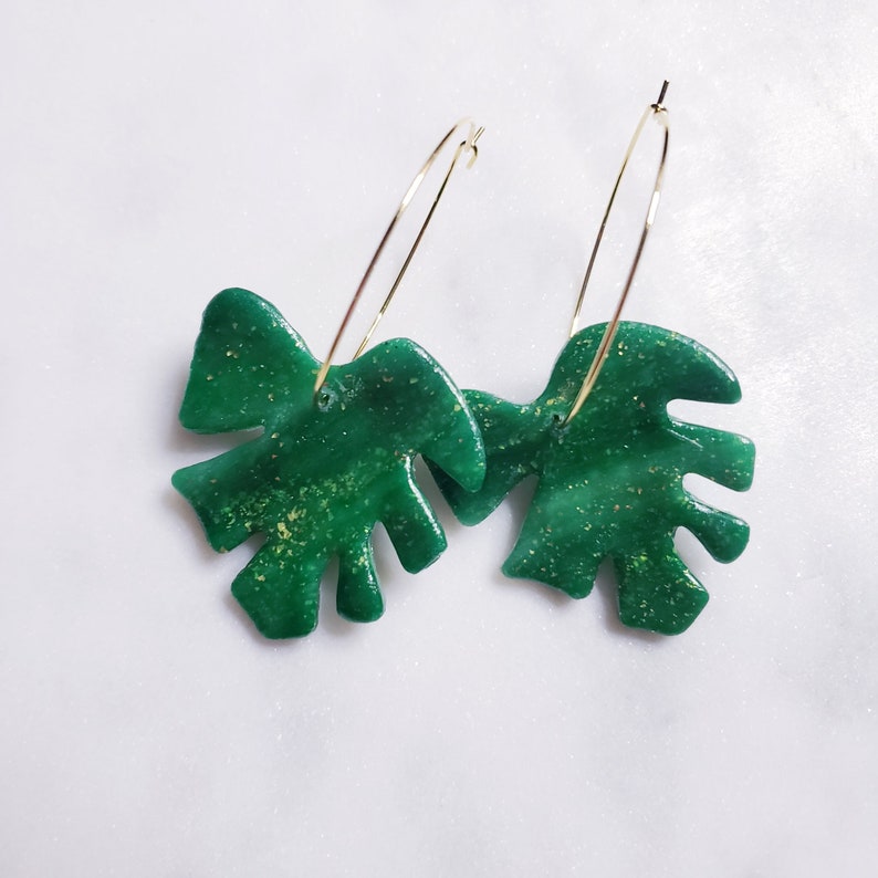 May include: A pair of gold hoop earrings with green glitter monstera leaf charms.