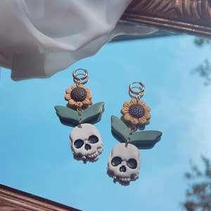 May include: A pair of dangle earrings featuring a skull, sunflower, and leaf design. The earrings have gold-colored hoops, gray skulls, yellow and brown sunflowers, and green leaves. The earrings are displayed on a reflective surface.