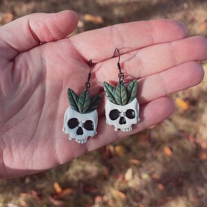 Puede incluir: Un par de pendientes &uacute;nicos con macetas blancas en forma de calavera con cuencas de ojos negras y hojas verdes. Los pendientes cuelgan de ganchos negros, mostrando una est&eacute;tica caprichosa y g&oacute;tica. Los pendientes se sostienen en una mano.