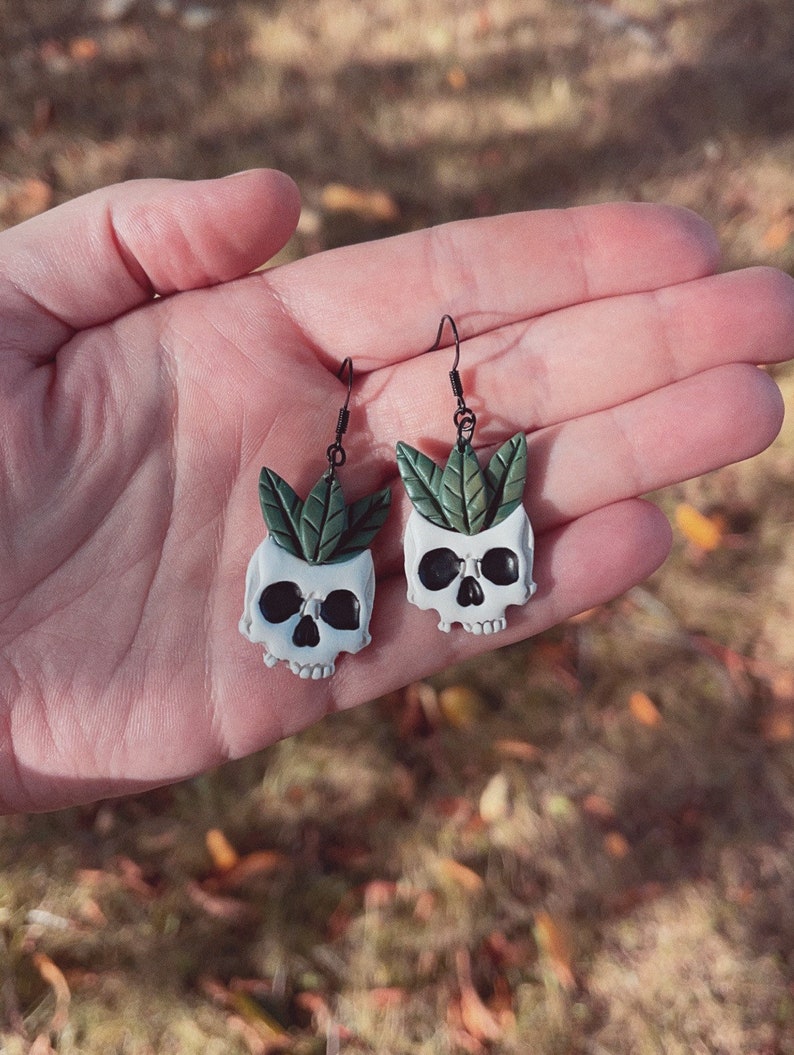 Puede incluir: Un par de pendientes &uacute;nicos con macetas blancas en forma de calavera con cuencas de ojos negras y hojas verdes. Los pendientes cuelgan de ganchos negros, mostrando una est&eacute;tica caprichosa y g&oacute;tica. Los pendientes se sostienen en una mano.