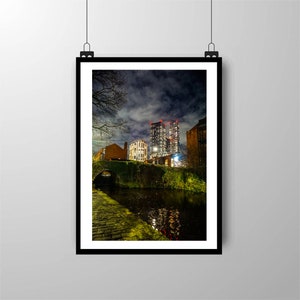 May include: A night view of a city with tall buildings lit up. A canal runs through the foreground with a stone bridge and lush greenery.