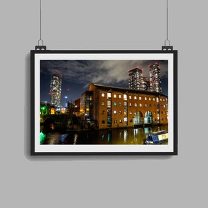 May include: A night view of a brick building with arched windows and a canal in front of it.  The building is lit up with warm yellow lights.  There are three tall buildings in the background, lit up with red lights.