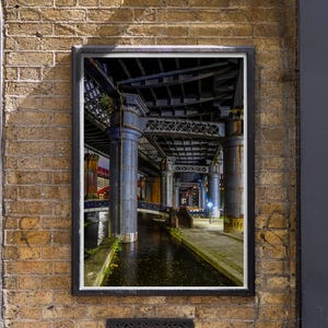 May include: A framed photograph of a dark, narrow canal running under a metal bridge. The bridge is made of steel beams and supports, and the canal is lined with brick walls. The photo is taken from a low angle, looking up at the bridge.