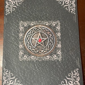 Könnte beinhalten: Ein dunkelgrauer Journal-Umschlag mit einem silbernen Pentagramm-Design und einem roten Juwel in der Mitte. Der Umschlag hat einen dekorativen Rand mit weißen floralen Mustern.