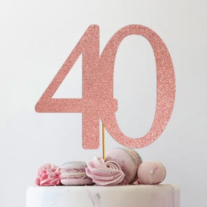 May include: A rose gold glitter "40" cake topper on a white cake with pink ribbon, macarons, and floral decorations. The cake has a marble effect. A gift box with gold polka dots is in the background.