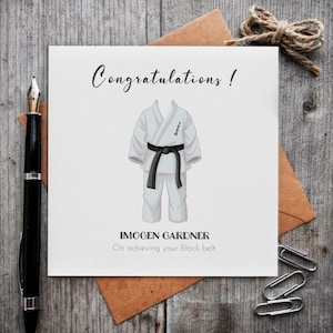 May include: A white card with the word "Congratulations!" in cursive. The card features a karate uniform illustration with a black belt. The text "IMOGEN GARDNER On achieving your Black belt" is also on the card. The card is on a wooden surface.