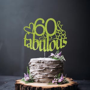 A white cake with a green glittery topper that says "60 & Fabulous" on it, surrounded by purple flowers.