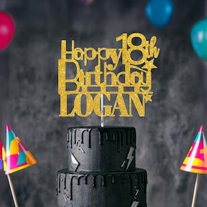 May include: A gold glitter cake topper featuring the text Happy 18th Birthday LOGAN with star accents. The topper is positioned on top of a two-tiered black cake with a drip design and lightning bolt decorations, set against a dark background.