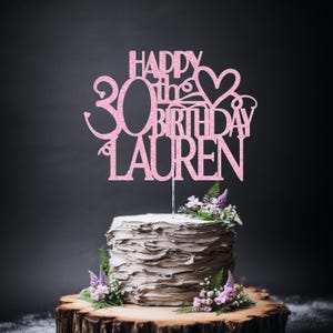 May include: A pink glitter cake topper featuring the text "HAPPY 30th BIRTHDAY LAUREN" with a heart shape integrated into the design. The topper is positioned on top of a textured, light-coloured cake decorated with small purple and green flowers.