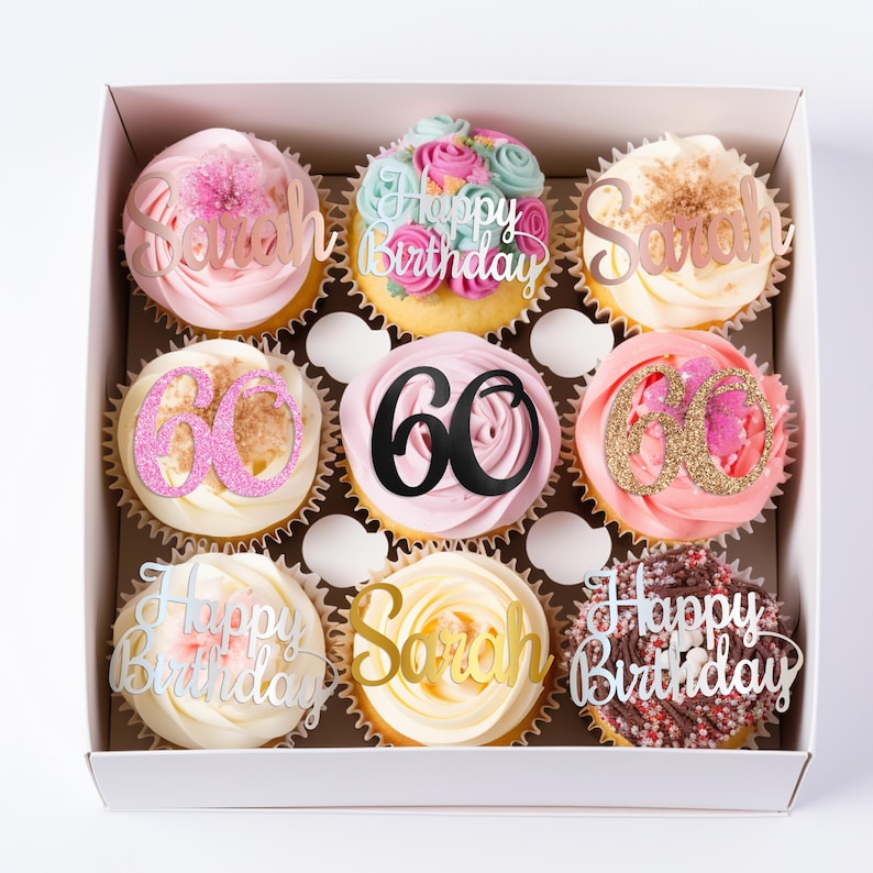 K&ouml;nnte beinhalten: Eine Schachtel mit neun Cupcakes mit verschiedenen Frosting-Farben und dekorativen Aufs&auml;tzen. Die Aufs&auml;tze zeigen den Namen Sarah in Ros&eacute;gold und Gold, die Zahl 60 in Pink-Glitzer, Schwarz und Gold-Glitzer sowie den Text Happy Birthday in Silber.