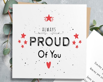 以下が含まれることがあります： 赤いハートと赤い星が描かれた、白地に黒文字で「Always Proud Of You」と書かれたカード。カードは茶色の表面の上に置かれている。