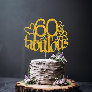 A white cake with a gold glittery topper that says "60 & Fabulous" on it, surrounded by purple flowers.