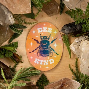May include: Oval sticker with a bee illustration and the words "BEE KIND" in white. The sticker has a gradient background of orange, yellow, and pink. The image is surrounded by crystals and greenery.