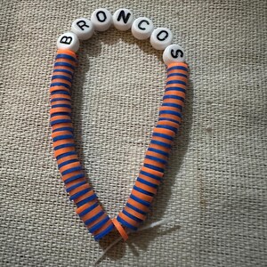 Könnte beinhalten: Ein Perlenarmband mit einer Reihe weißer runder Perlen, auf denen der schwarze Text BRONCOS aufgedruckt ist, gefolgt von einem langen Abschnitt aus abwechselnd orangefarbenen und blauen flachen runden Scheibenperlen in einem Streifenmuster.