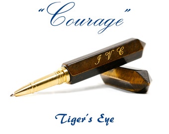 May include: A gold and brown pen with the inscription "JVC" on the barrel. The pen is made of tiger's eye stone and has a gold tip. The pen is lying on a white surface with the words "Courage" and "Tiger's Eye" written in blue cursive.