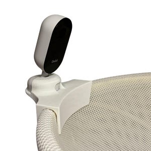 May include: A white baby monitor camera with a black lens is attached to a white bracket, clipped to the edge of a white mesh crib. The camera has the brand name "Owlet" printed on it.