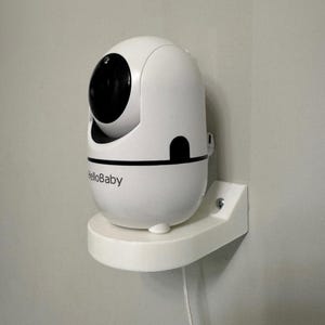 Puede incluir: Un monitor para bebés HelloBaby blanco montado en un soporte de pared blanco. El monitor tiene una lente negra y un diseño redondeado. El texto "HelloBaby" está impreso en la parte frontal del monitor.