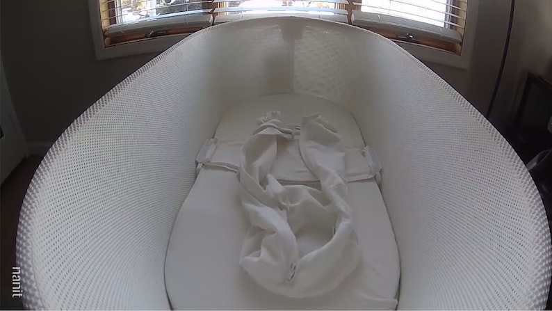 May include: A white mesh bassinet with a white fitted sheet and a white blanket inside. The bassinet is made by the company "nanit".