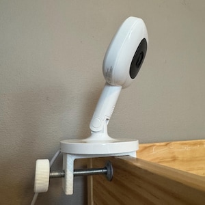 May include: A white baby monitor camera with a black lens is clamped to a wooden crib rail. The camera has a rounded head and a flexible neck. A white adjustment knob is visible on the clamp.