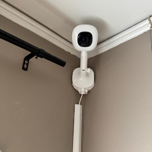 May include: A white security camera mounted in the corner of a room. The device features a rounded, square-shaped camera head with a black lens, attached to a vertical stand and a wall-mounted base. A white power cable runs downward into a white cover.